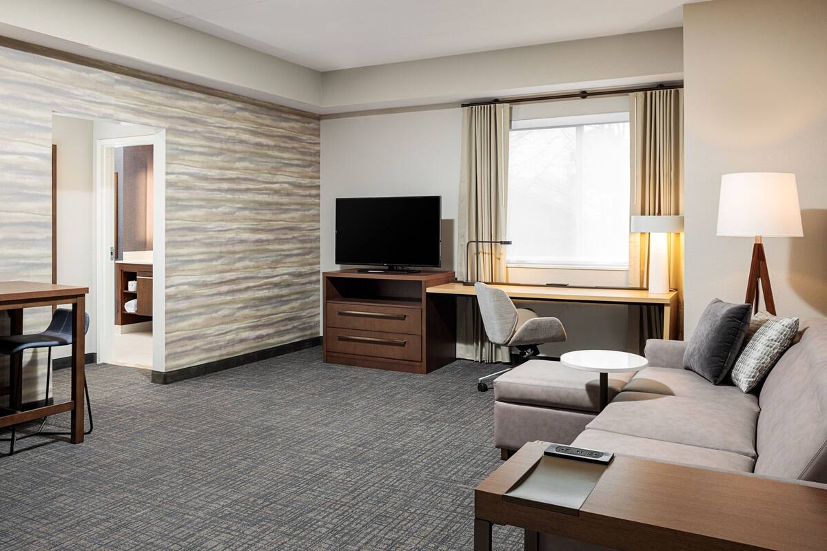 A hotel room with a flat-screen TV, a sofa, a desk with a chair, and a lamp.