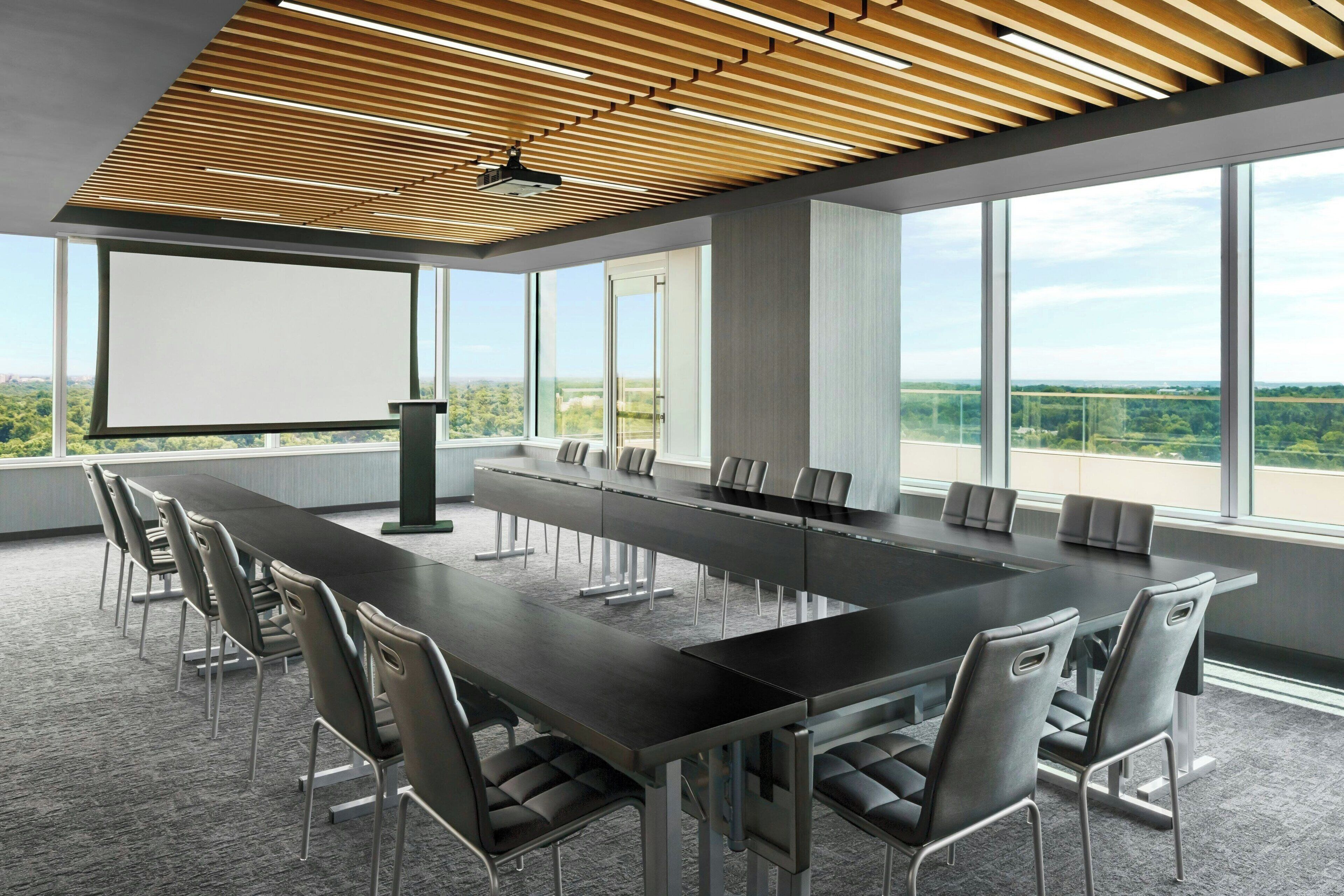 Ein Konferenzraum mit einem langen Tisch, Stühlen, einer Projektionsleinwand und großen Fenstern mit Blick auf die Landschaft.