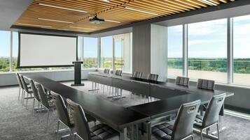 Ein Konferenzraum mit einem langen Tisch, Stühlen, einer Projektionsleinwand und großen Fenstern mit Blick auf die Landschaft.