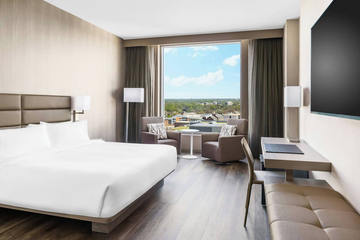 Een moderne hotelkamer met een groot bed, een zithoek met een tafel, en een uitzicht op de skyline door grote ramen.