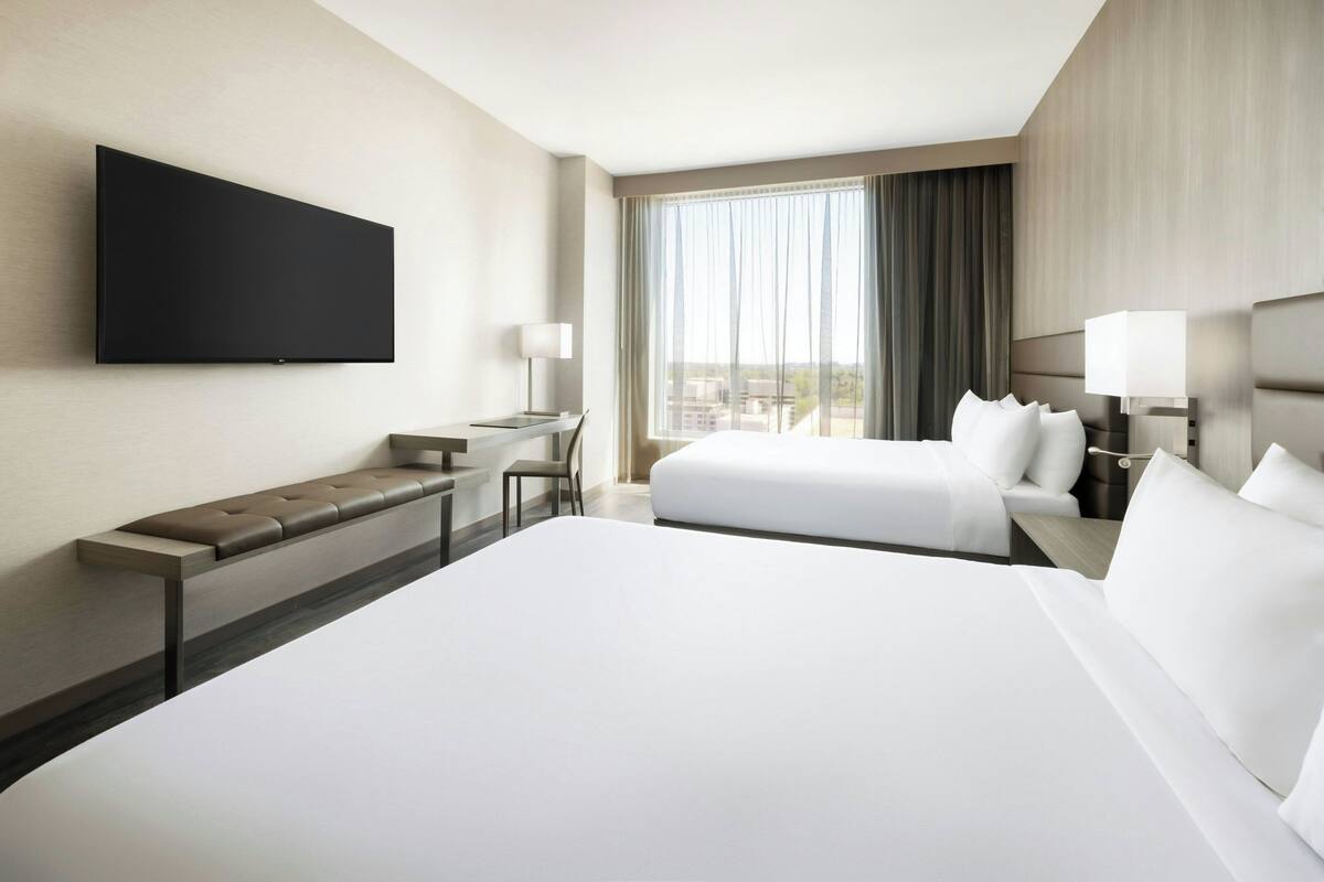 Een hotelkamer met een groot bed, een kleiner bed, een bureau met stoel, een flatscreen televisie aan de muur en een raam met gordijnen.