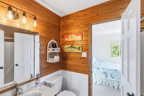 Un baño con paredes de madera, un inodoro blanco, un lavabo con espejo y un estante con artículos de aseo.