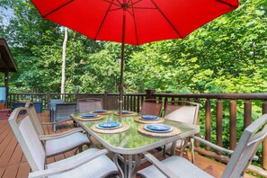 A deck with a table and chairs, surrounded by a wooden railing, set in a lush green environment. - Great Smokies Adventure Cabin w/Hot Tub and Deck! (Sevierville)