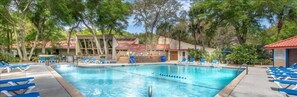 Una piscina con sillas reclinables azules, rodeada de árboles y un edificio de techo rojo.