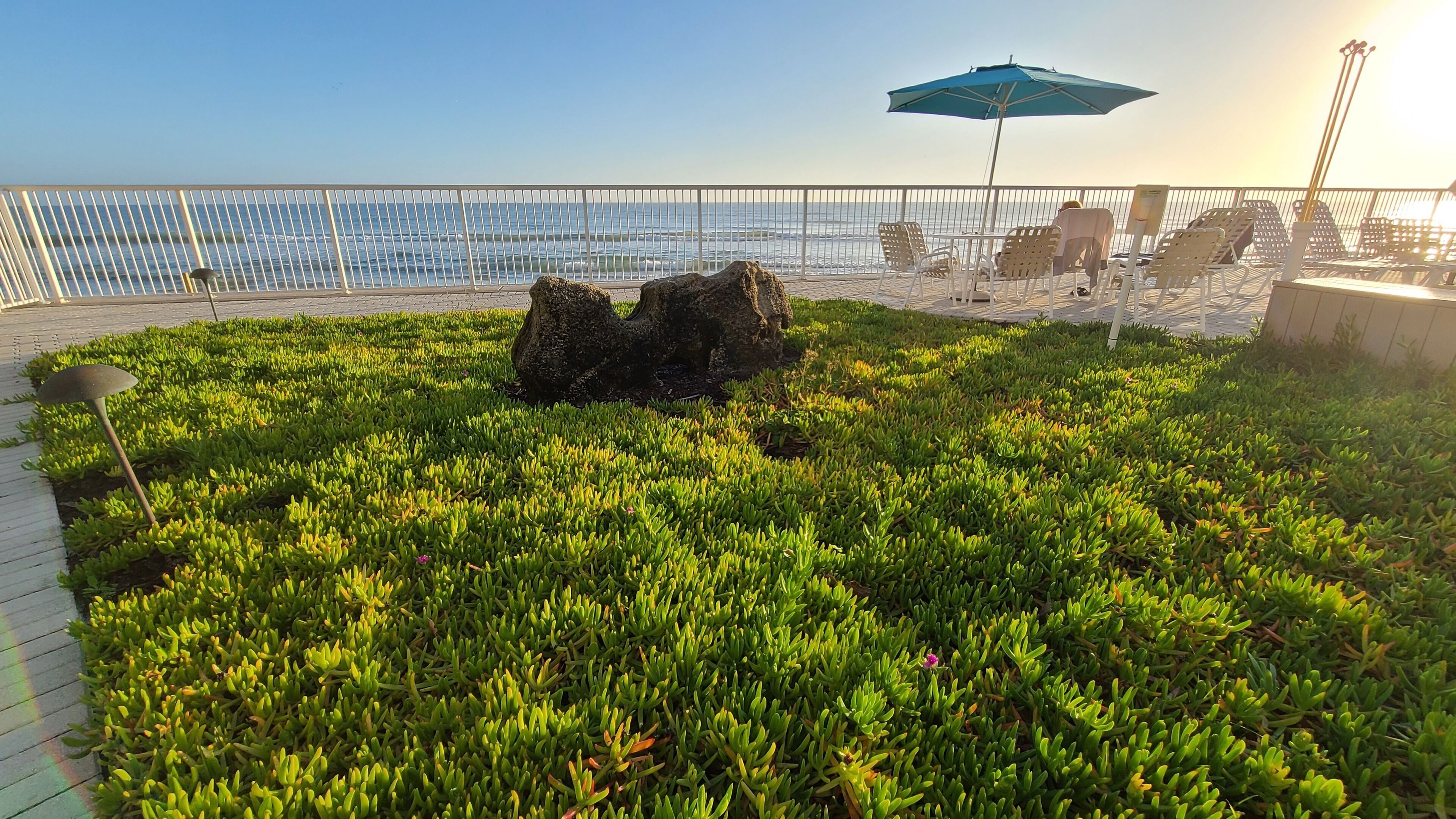 Un giardino pensile con vista sul mare, che include una formazione rocciosa, un ombrellone blu e posti a sedere all'aperto.