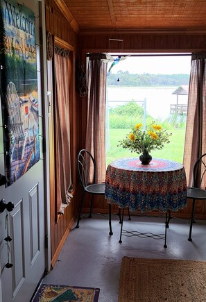 Une pièce avec vue sur un lac, une table ronde recouverte d’une nappe à motifs, deux chaises et un vase rempli de fleurs.