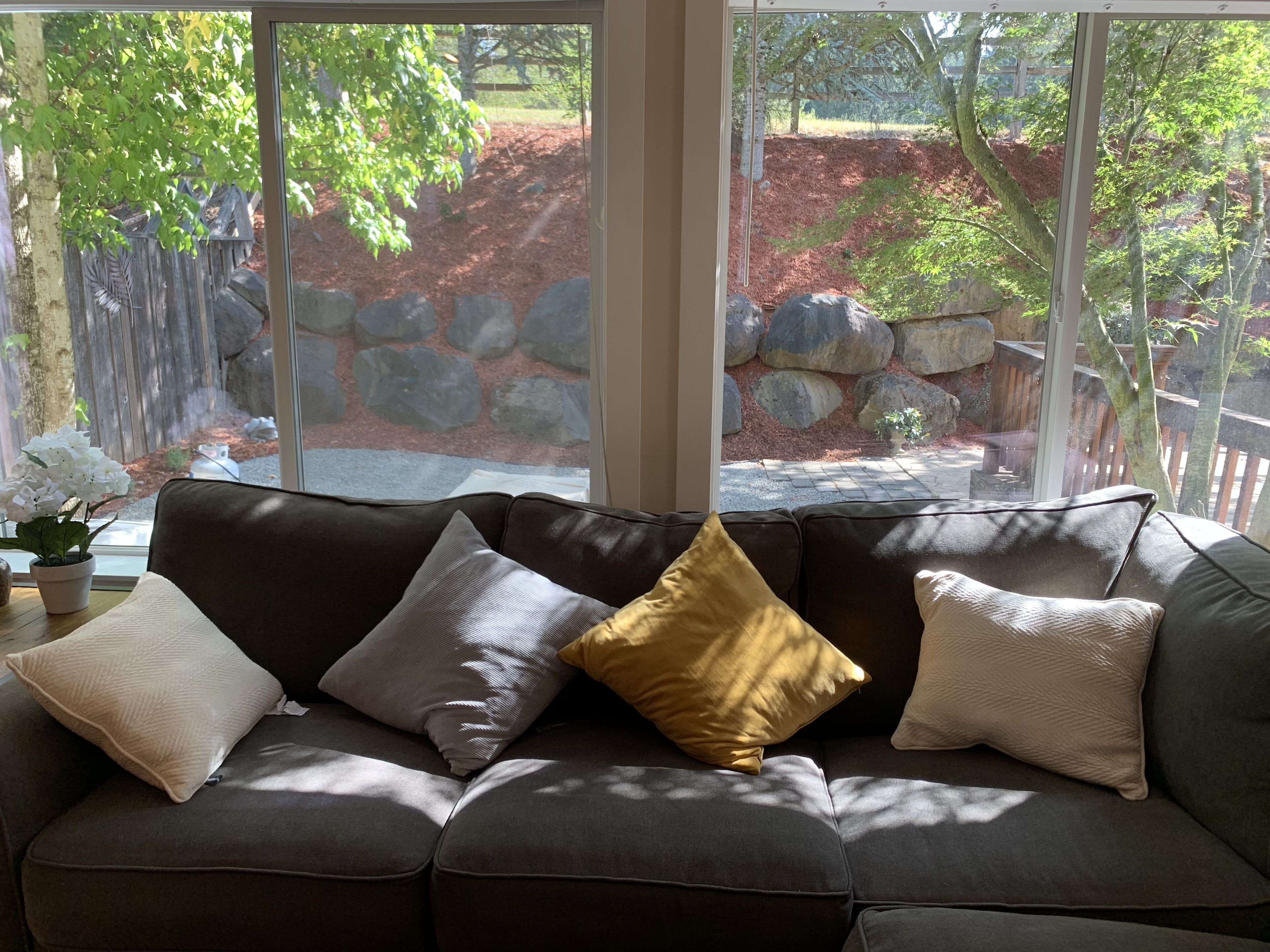Un sofá marrón con tres cojines en una habitación con amplios ventanales que dan a un área ajardinada con rocas y árboles.