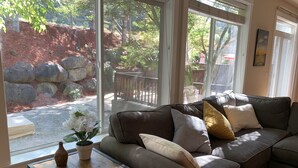 A modern living room with a brown leather sofa, a small wooden side table, and a large window offering a view of a stone wall and greenery outside. - Miller Hill Sanctuary near Intel w/ BBQ & Firepit (Aloha)