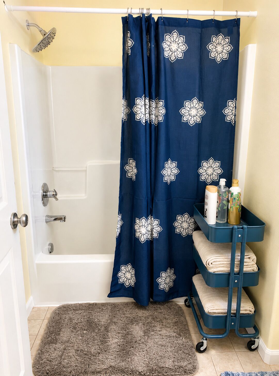A bathroom with a shower, a bathtub, a blue shower curtain with a floral pattern, a towel rack with towels, and a small cart with toiletries.