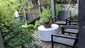Un patio in legno con un tavolo rotondo e due sedie, immerso nel verde e delimitato da un muro in pietra.