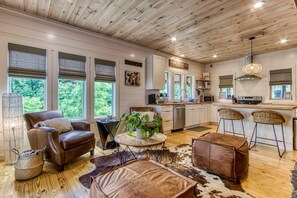 A spacious living room with a leather sofa, a round coffee table, and a cowhide rug. The kitchen features white cabinets, a stainless steel range hood, and a bar area with stools.