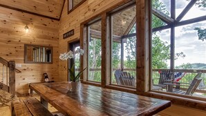 A wooden interior with a large table, benches, and large windows offering a view of outdoor seating and greenery.