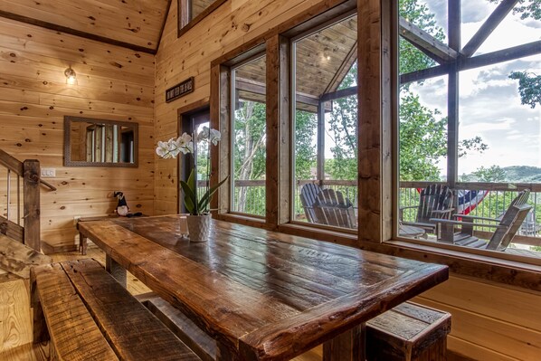 A wooden interior with a large table, benches, and large windows offering a view of outdoor seating and greenery.