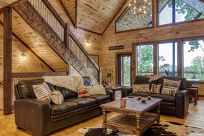 A spacious living room with a wooden staircase, leather sofas, and a large window offering a view of the outdoors.