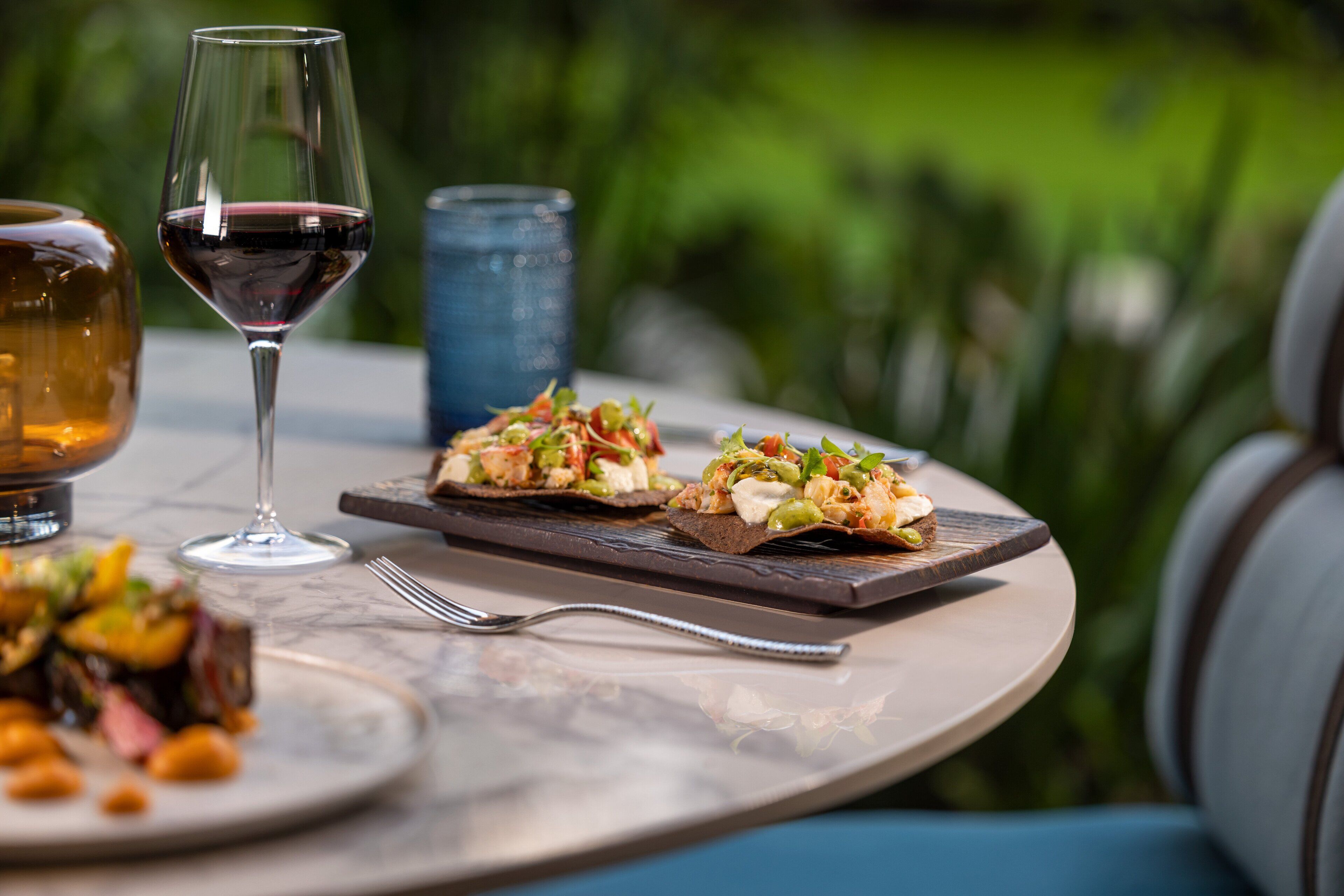 A dining table with a glass of red wine, a blue glass, and two plates of food.