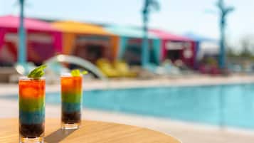 Dos bebidas en capas sobre una mesa de madera al lado de una piscina, con un edificio colorido de fondo.