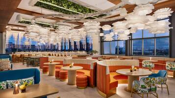A modern lounge with tiered seating, hanging floral light fixtures, and large windows offering city views.