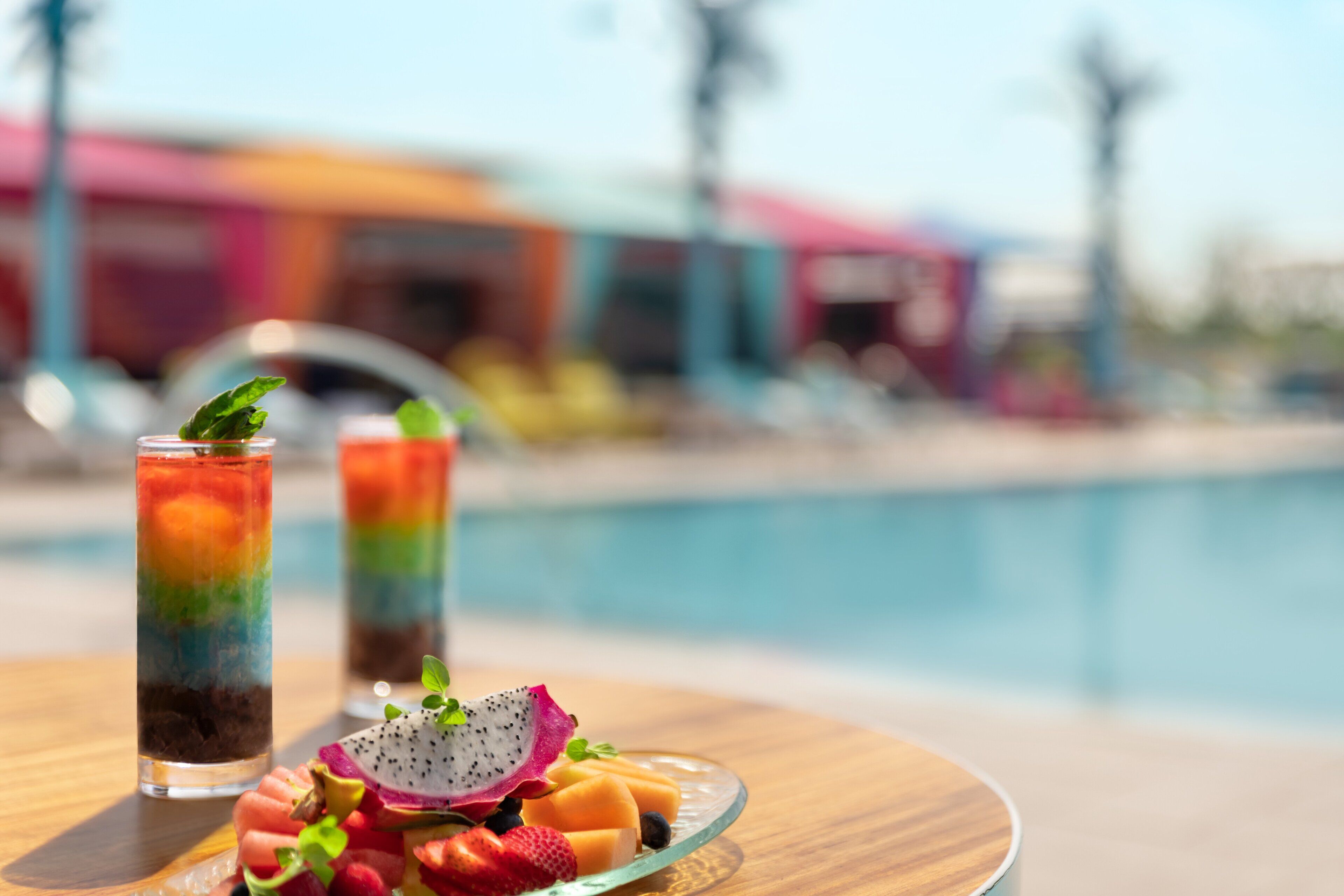 Deux grands verres contenant des boissons à plusieurs couches, garnies de fruits, sont posés sur une table en bois au bord d’une piscine, avec un bâtiment coloré en arrière-plan.