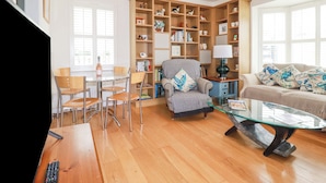 Un salon comprenant une table basse en verre, un canapé agrémenté de coussins décoratifs, une bibliothèque et un coin repas avec des chaises et une table.