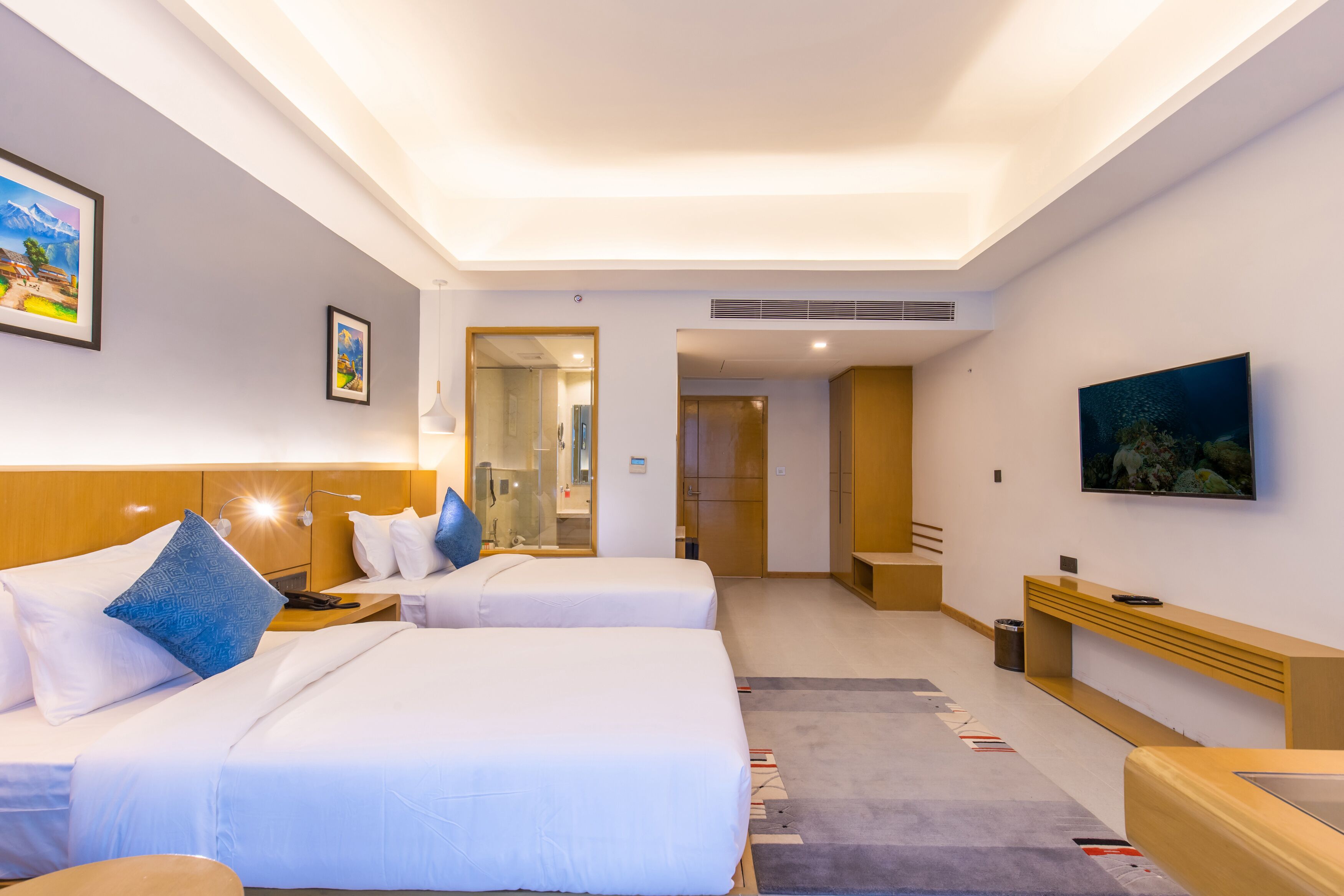 Habitación de hotel con dos camas, televisión, escritorio y baño visible a través de una puerta de cristal.