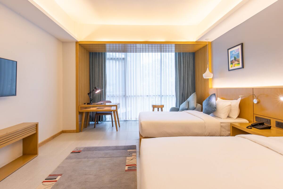 A hotel room with two beds, a desk, a chair, a TV, and a window with curtains.