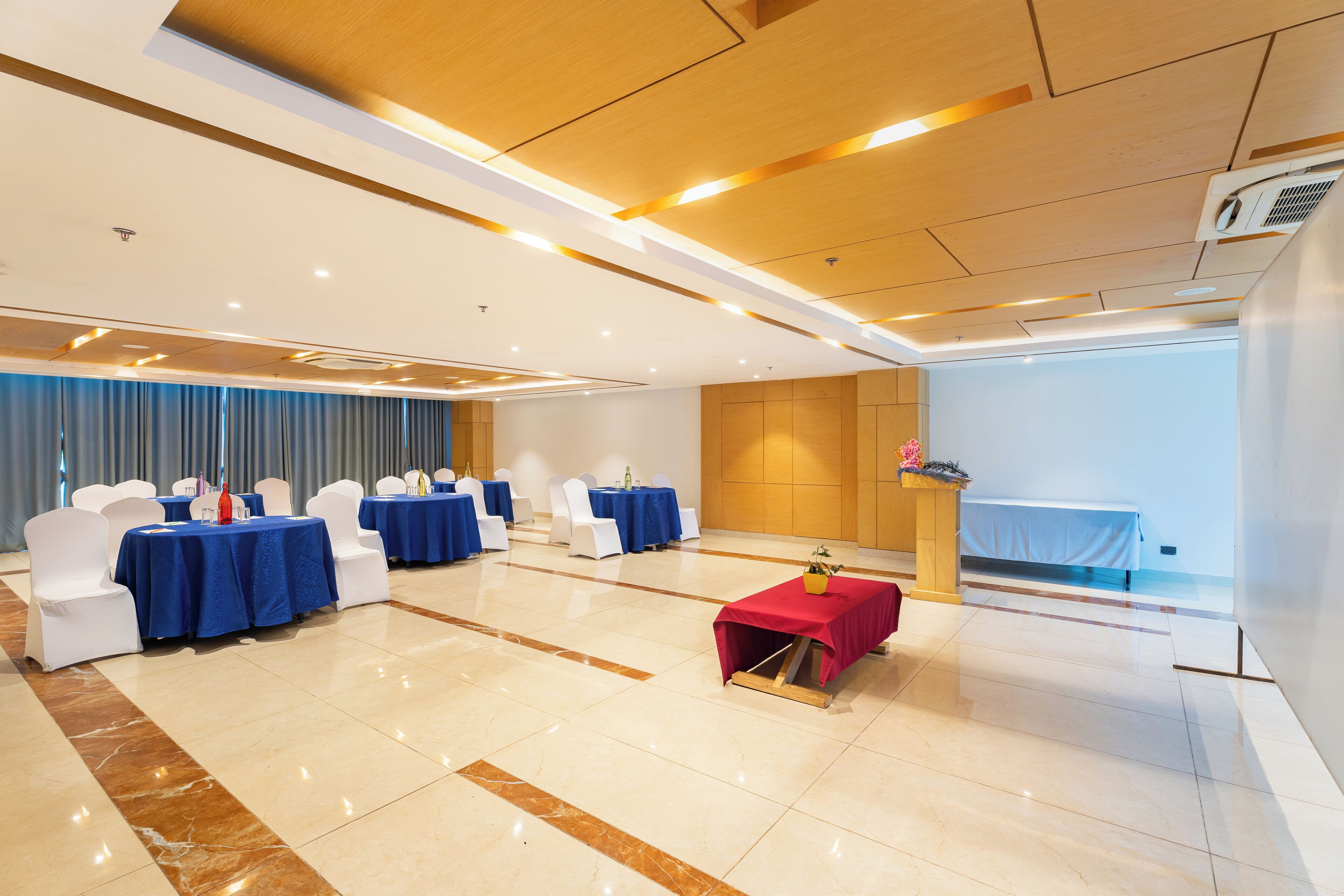 A conference room with round tables, white chairs, a red table with a vase, and a bed in the background.