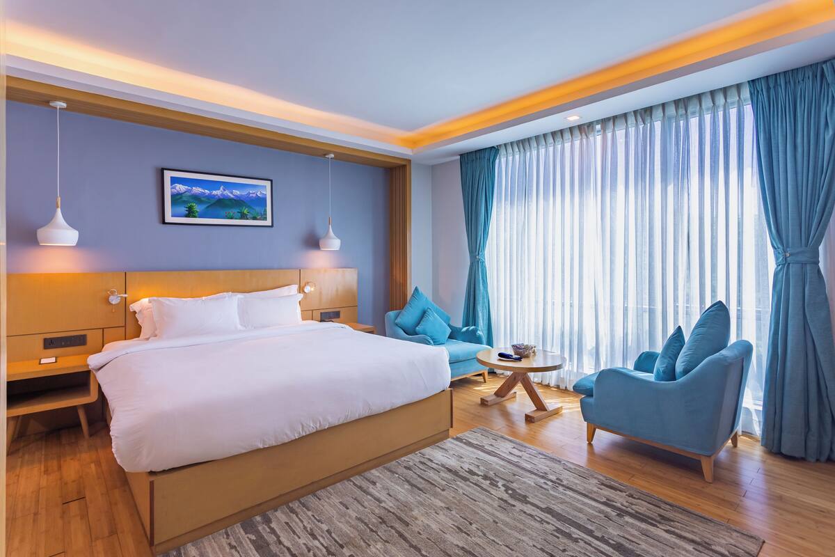 A modern hotel room with a large bed, a blue armchair, a small round table, and a painting on the wall.