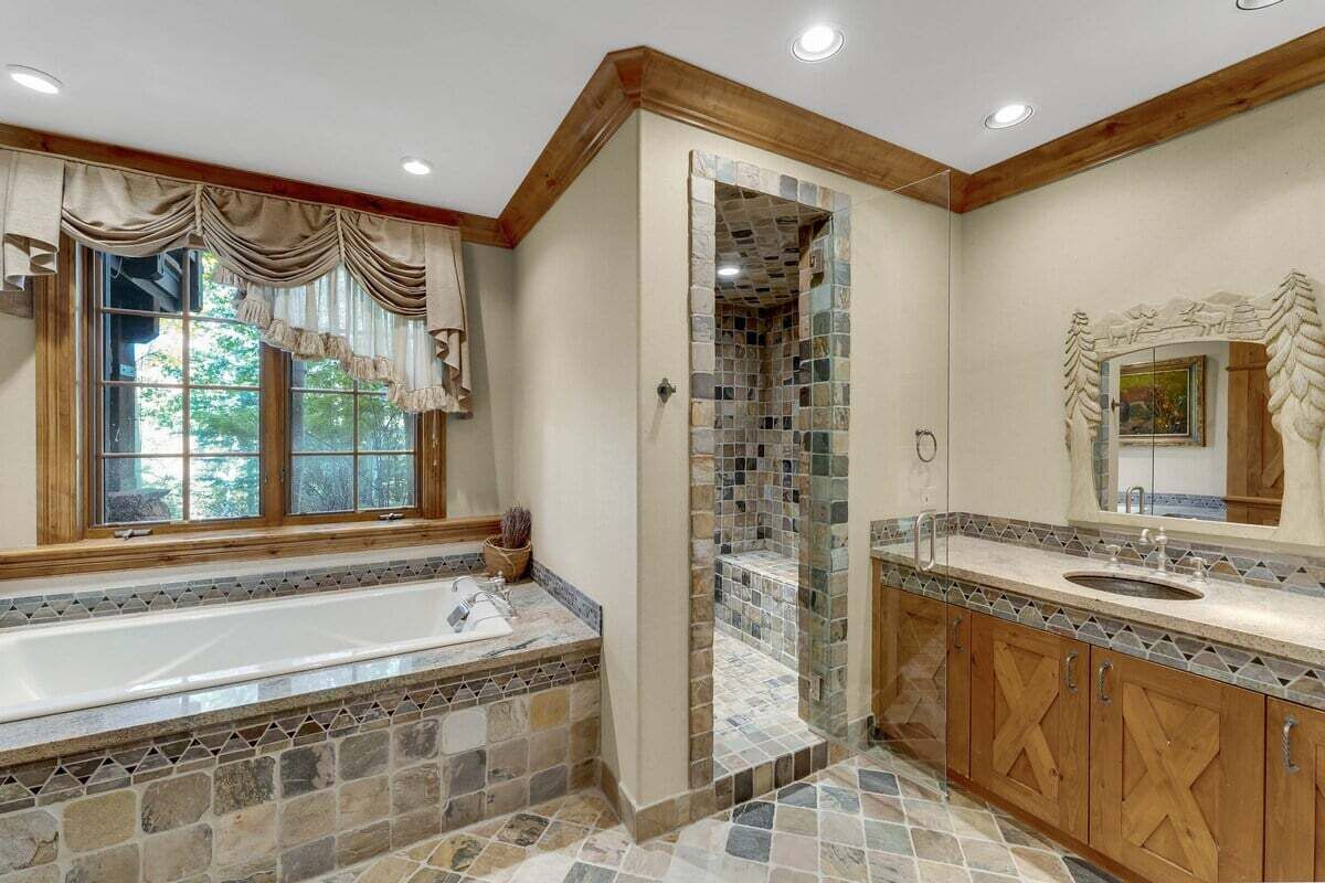 A bathroom with a stone-tiled shower, a large bathtub, wooden cabinets, and a window with curtains.