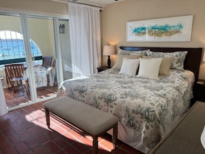 Quarto com cama, um banquinho, mesa de cabeceira e vista para o mar através de uma porta deslizante.