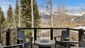 A wooden deck with Adirondack chairs and a stone fire pit, overlooking a mountainous landscape with snow-capped peaks.
