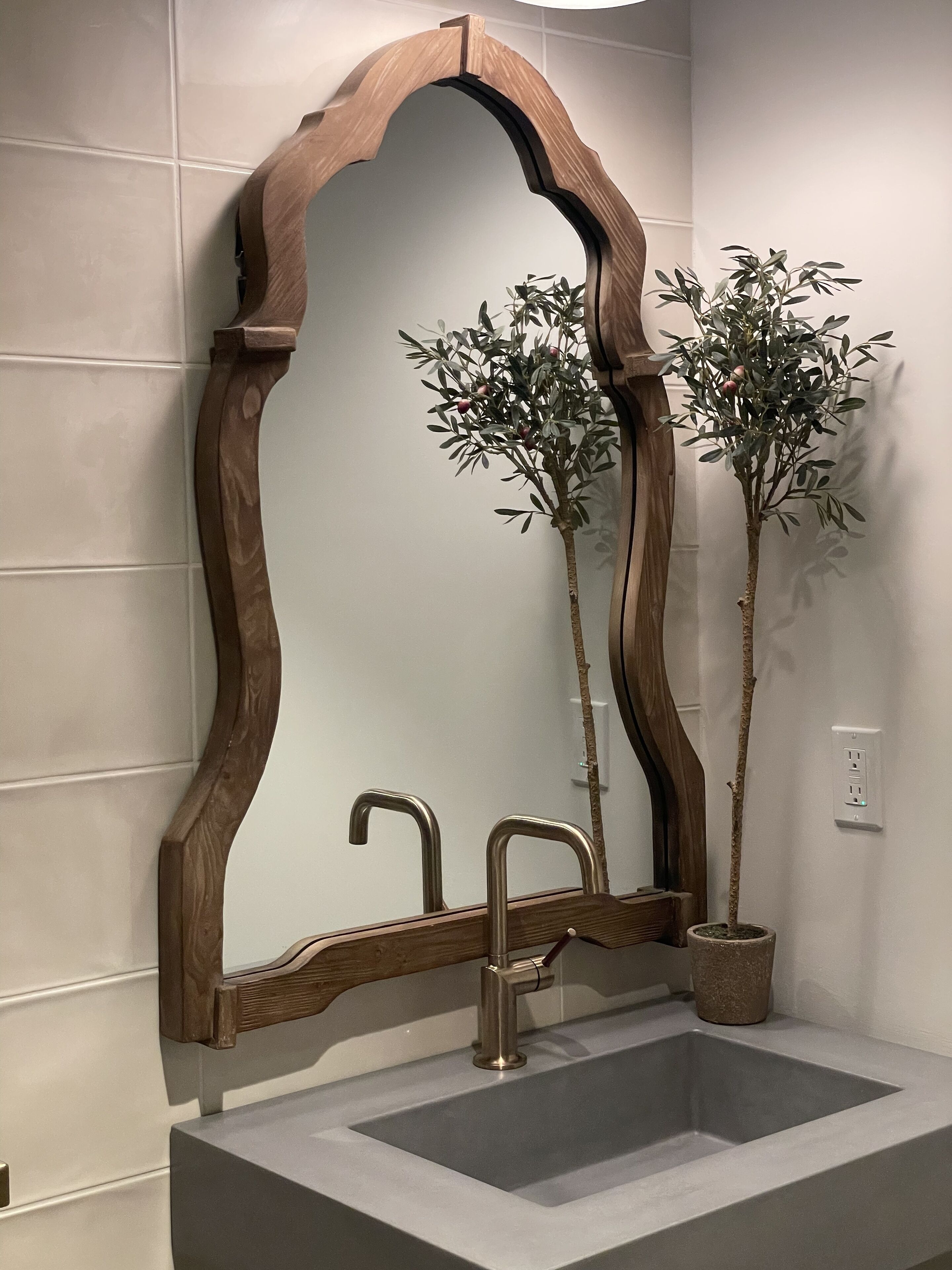 Um banheiro com espelho de moldura de madeira, pia e duas plantas em vasos.