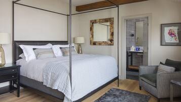 A four-poster bed with a headboard, a nightstand with a lamp, a gray sofa, and a framed artwork on the wall.