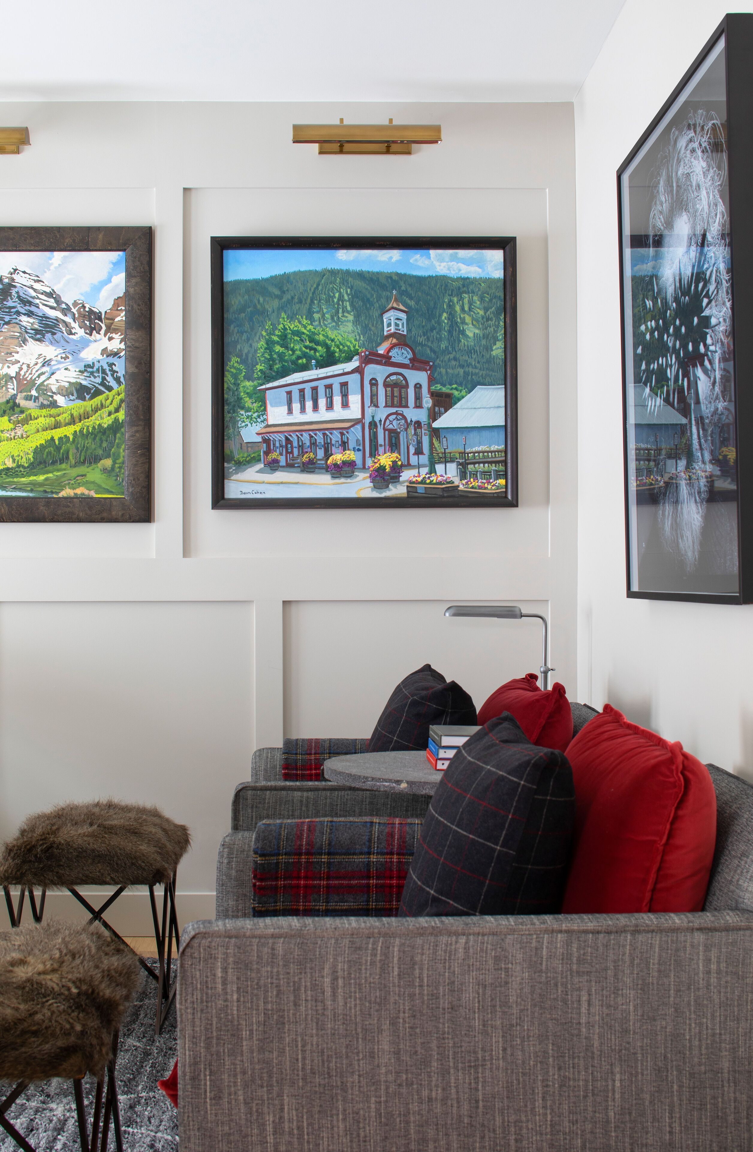 Una sala de estar con un sofá gris, cojines rojos y de cuadros, una mesita y un cuadro enmarcado de una casa con una torre del reloj.