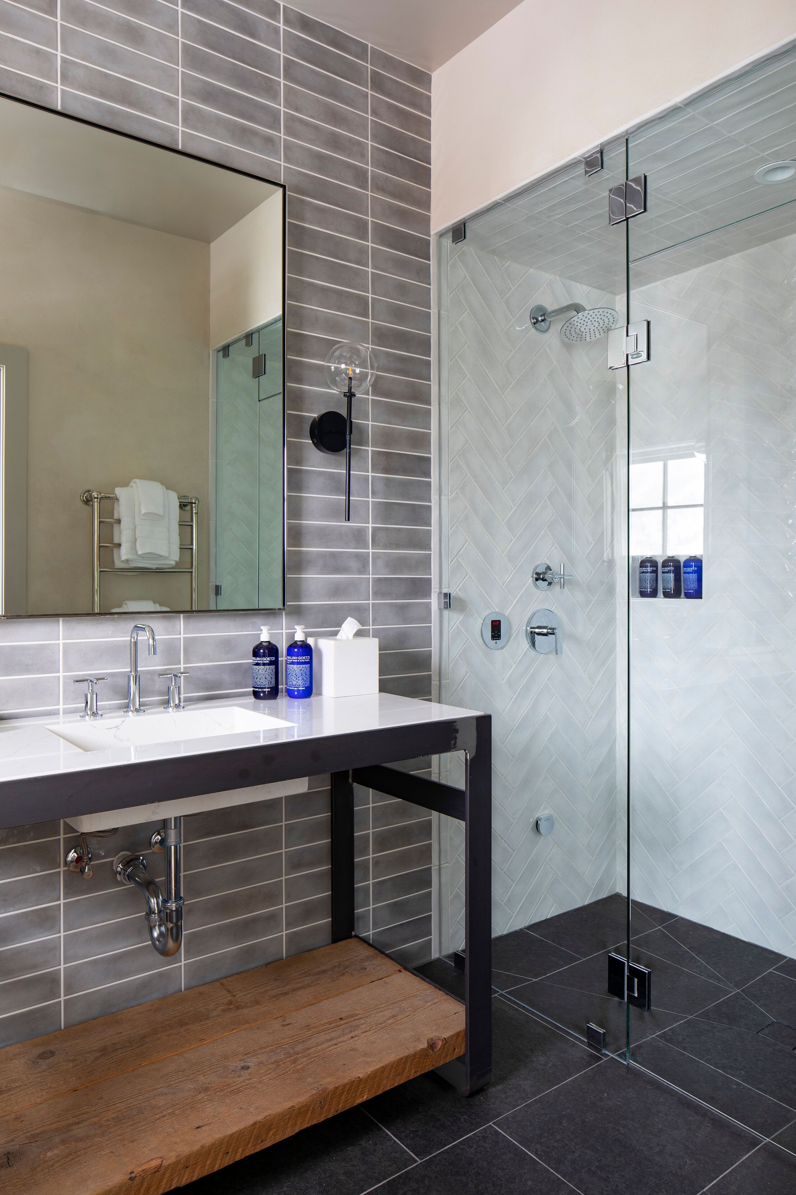 A modern bathroom with a large mirror, two sinks, a shower enclosure, and a wooden bench.