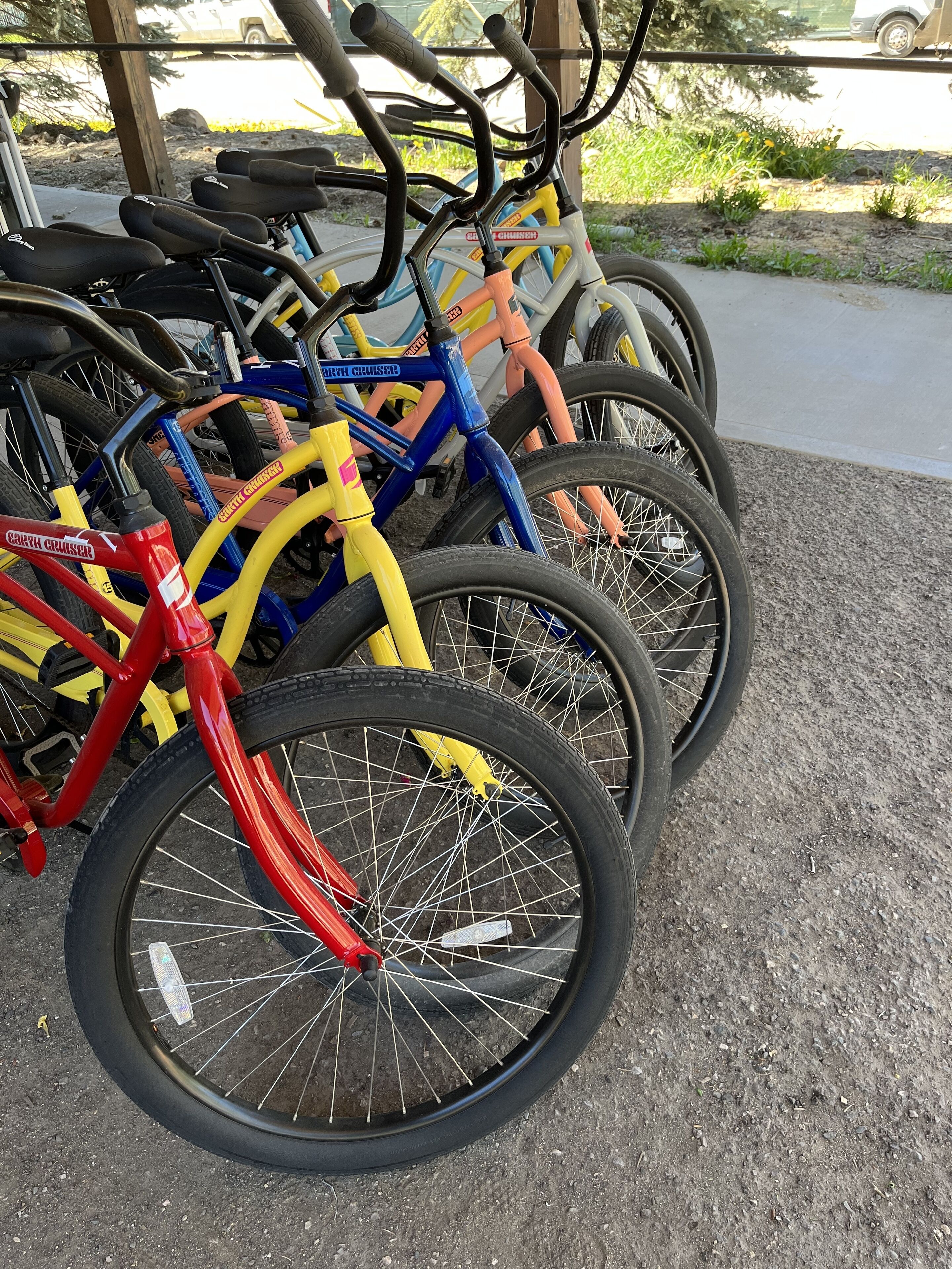 A row of bicycles parked outdoors, with a gravel surface and a wooden structure in the background.