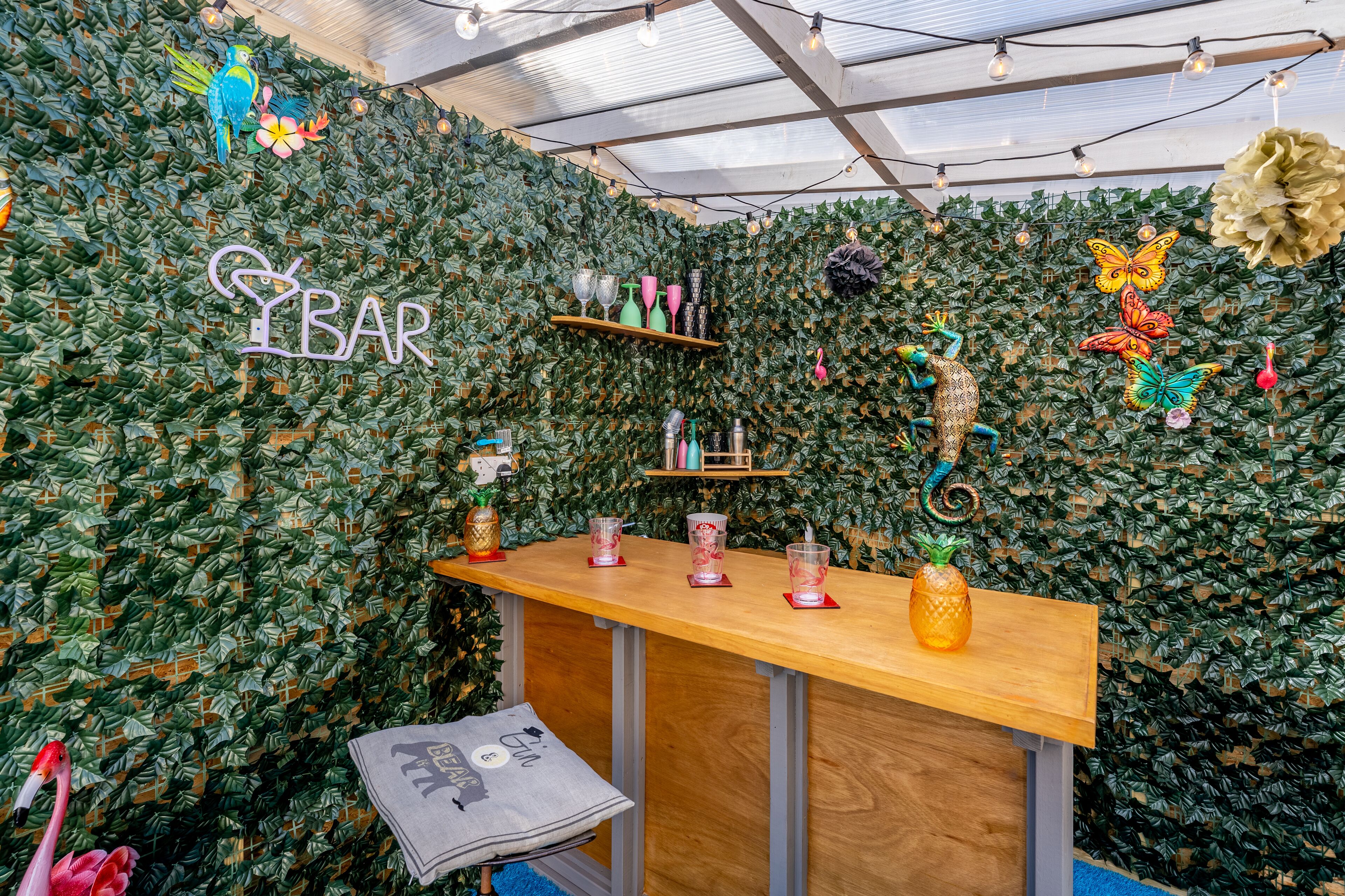 Zona de bar con mostrador de madera, estantes con vasos y elementos decorativos como una piña y flamencos, todo sobre un fondo de pared con hojas.