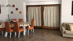 Zona pranzo con tavolo imbandito per quattro persone, sedie arancioni, un lampadario e una porta a vetri scorrevoli con tende.