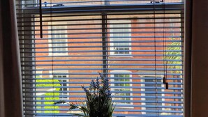 A window with horizontal blinds, a potted plant on the sill, and a view of a brick building with a chimney. - Charming Mews House in the Heart of Lymington (Lymington)