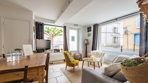 A living room with a wooden dining table, a sofa, a TV, and a window with a view of the street.
