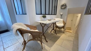 A room with a white tiled floor, two wicker chairs with white fur throws, a marble table, and a window with vertical blinds.