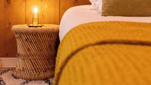 A bed with white and green pillows, a yellow blanket, and a lit lamp on a woven stool.
