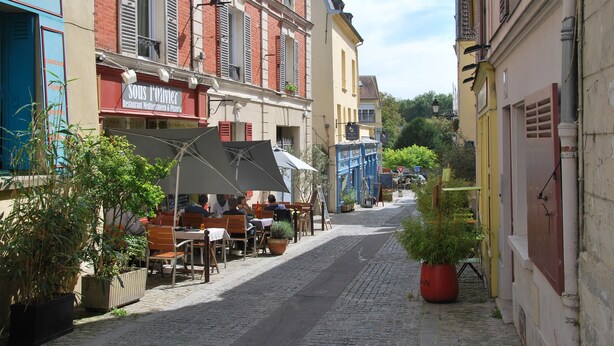 A cobblestone street lined with cafes and restaurants, featuring outdoor seating and potted plants.