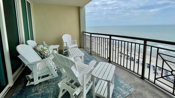 Un balcon avec des fauteuils Adirondack blancs et une table, donnant sur une plage parsemée de parasols et fréquentée par des gens.