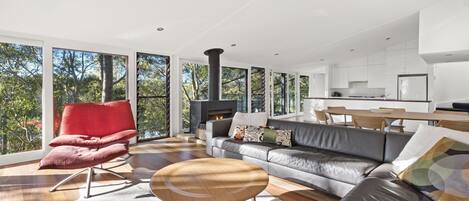 A modern living room with a fireplace, a red chair, a sofa, and a dining area with a kitchen in the background.