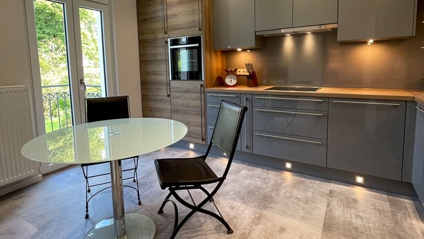 A modern kitchen with a round glass table and a black chair, wooden cabinets, and a view of greenery through large windows. - Saar-Apartments (Merzig)