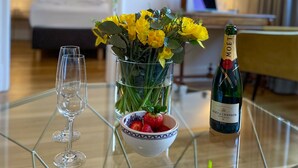 A hotel room with a bed, a desk, and a glass table with a bottle of champagne, a bowl of strawberries, and a vase of yellow flowers. - Saar-Apartments (Merzig)
