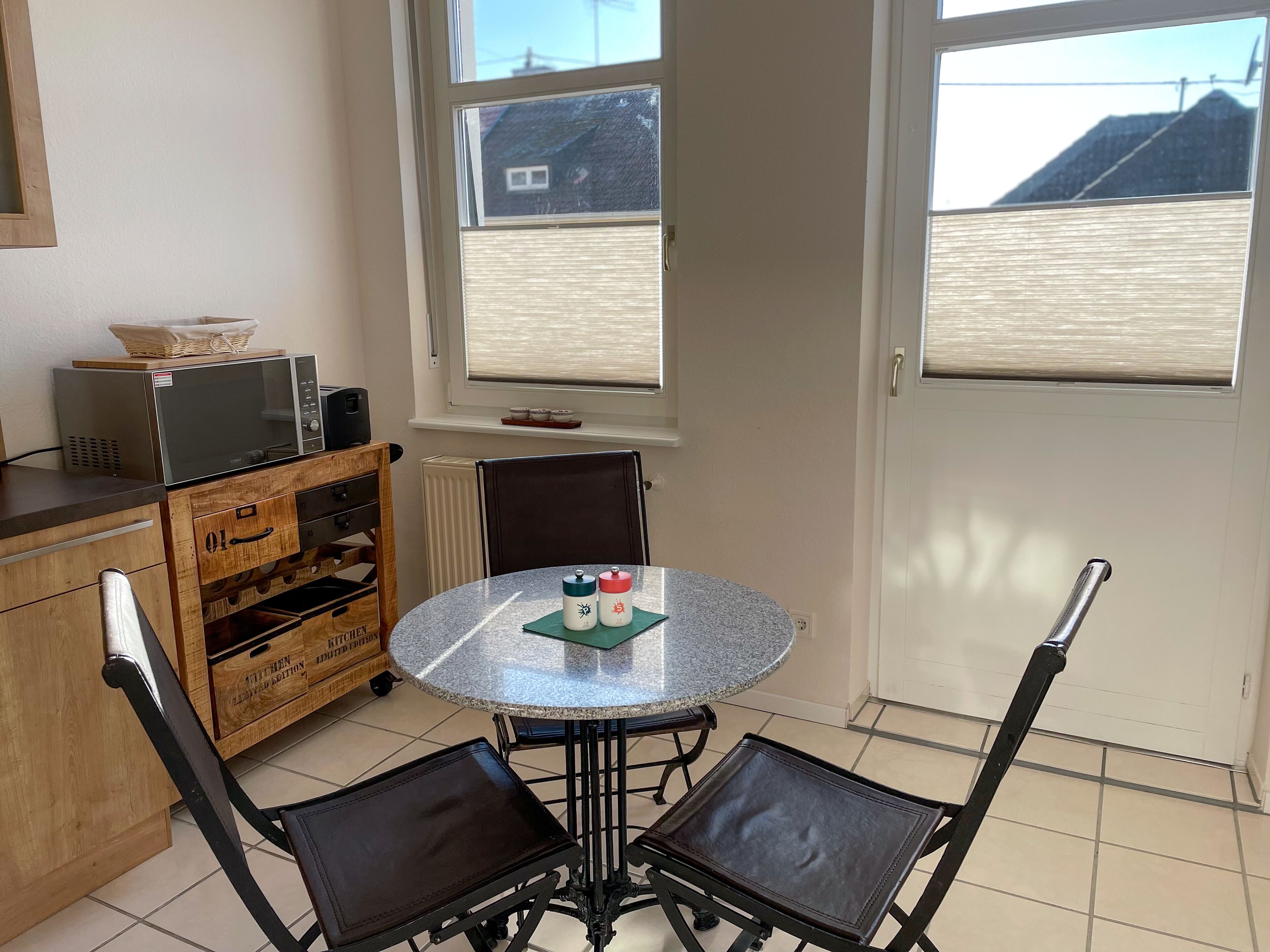 Una cocina con una mesa redonda, dos sillas, un microondas, un armario de madera y una ventana con vista a un edificio.