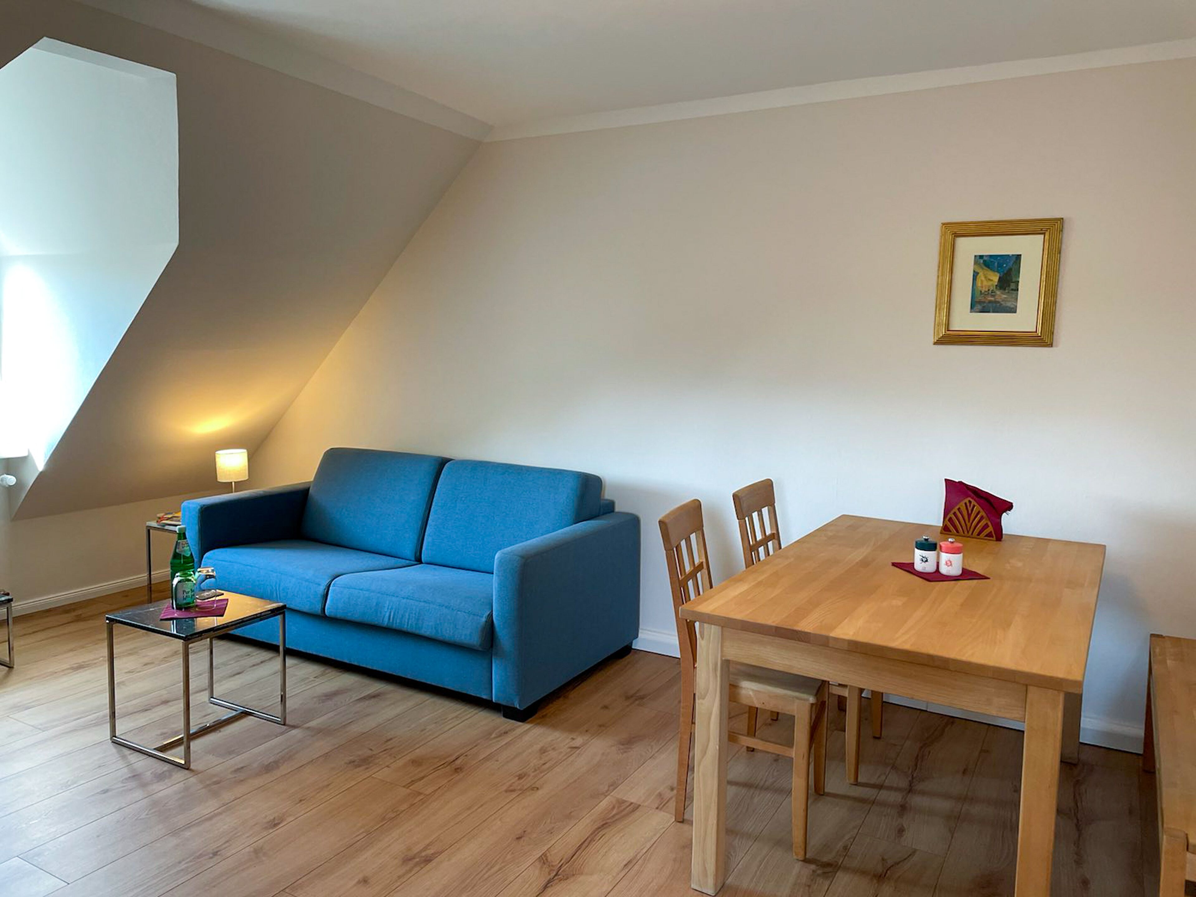 Una sala de estar con un sofá azul, una mesa de comedor de madera y una pequeña mesita de noche con una lámpara.