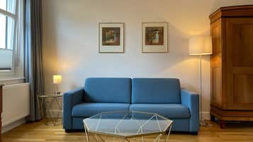 A living room with a blue sofa, a glass coffee table, two framed artworks on the wall, a floor lamp, and a wooden cabinet.