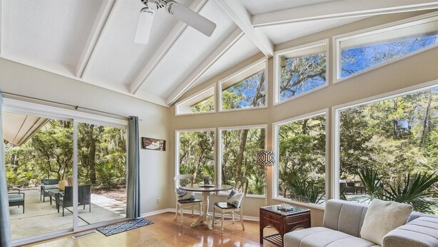 Un vaste espace de vie avec une grande fenêtre donnant sur un jardin, un ventilateur de plafond et un coin salon confortable avec un canapé et des fauteuils.